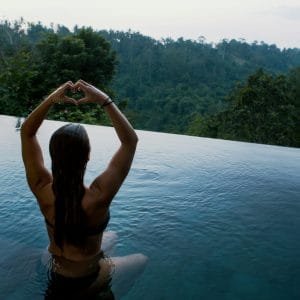 woman in infinity pool making heart hand gesture facing green leafed trees